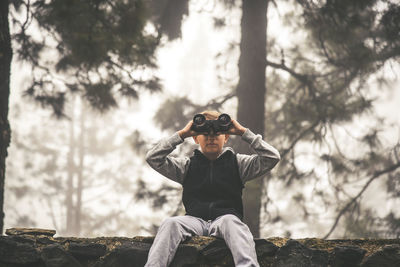 Boy looking through binoculars while sitting on retaining wall at forest