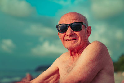 Portrait of shirtless man wearing sunglasses sitting at beach against sky