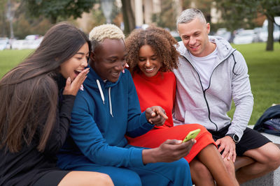 Smiling friends looking at phone sitting on bench at park