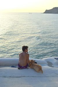 Rear view of shirtless man sitting with dog on scala dei turchi by sea during sunset