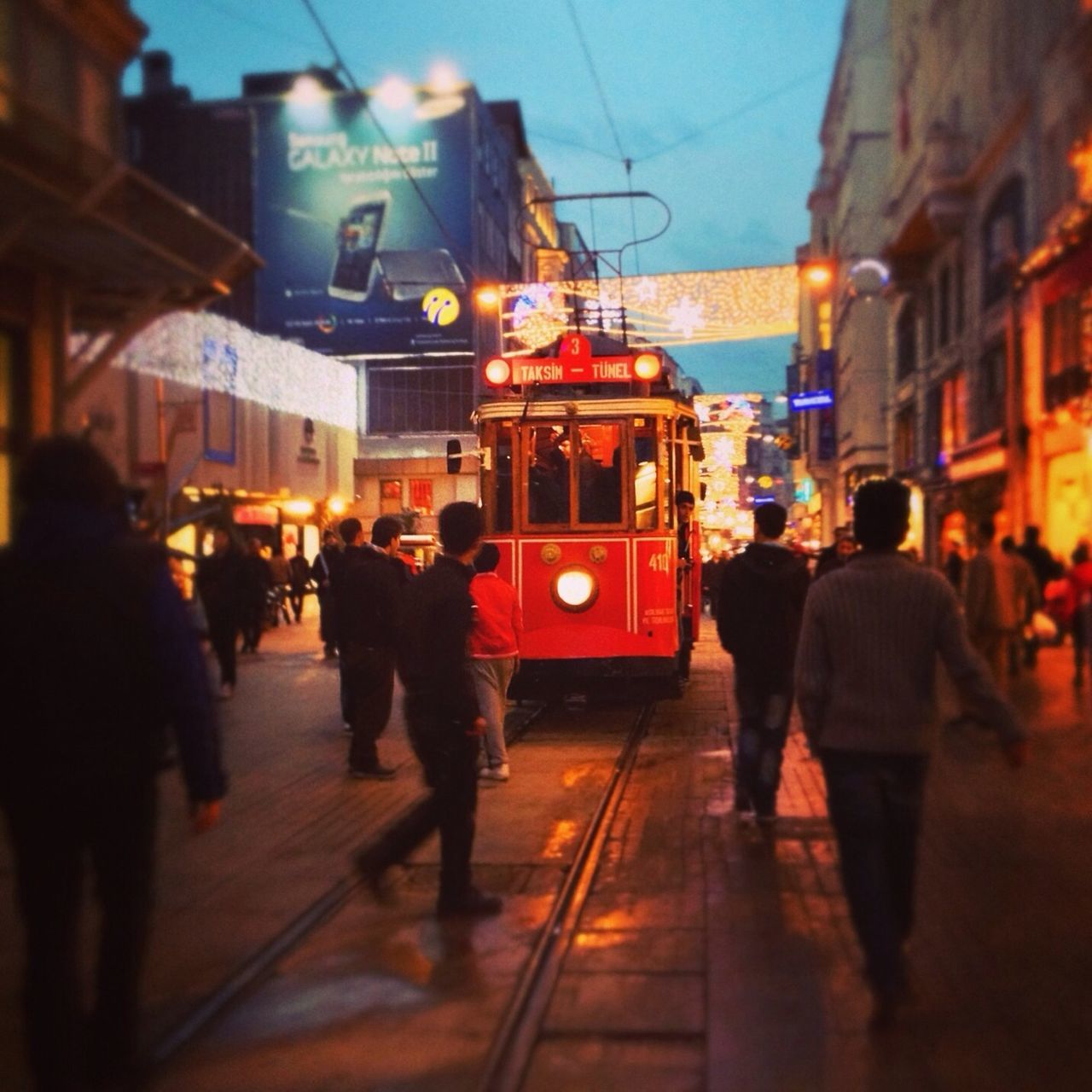 building exterior, architecture, built structure, city, city life, men, street, person, walking, transportation, large group of people, lifestyles, city street, illuminated, railroad track, on the move, mode of transport, public transportation, rear view