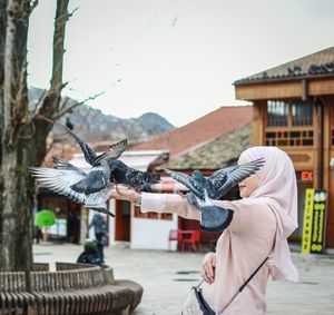 Side view of young woman feeding birds while standing on city street