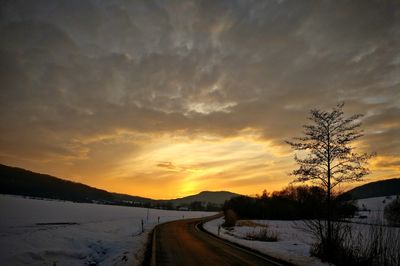 Road by snow covered trees against sky during sunset