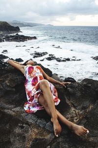 Midsection of man relaxing on rock at beach against sky