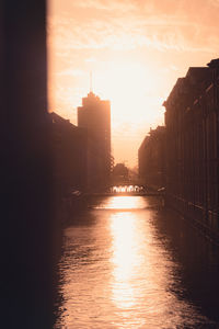 Silhouette buildings by river against sky during sunset