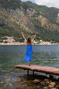 A girl in a dress stands with her hands up on the pier of the bay of kotor in montenegro.