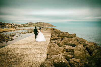 Rear view of young couple walking at beach against cloudy sky