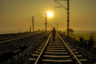 Man walking on railroad track against sky during sunset