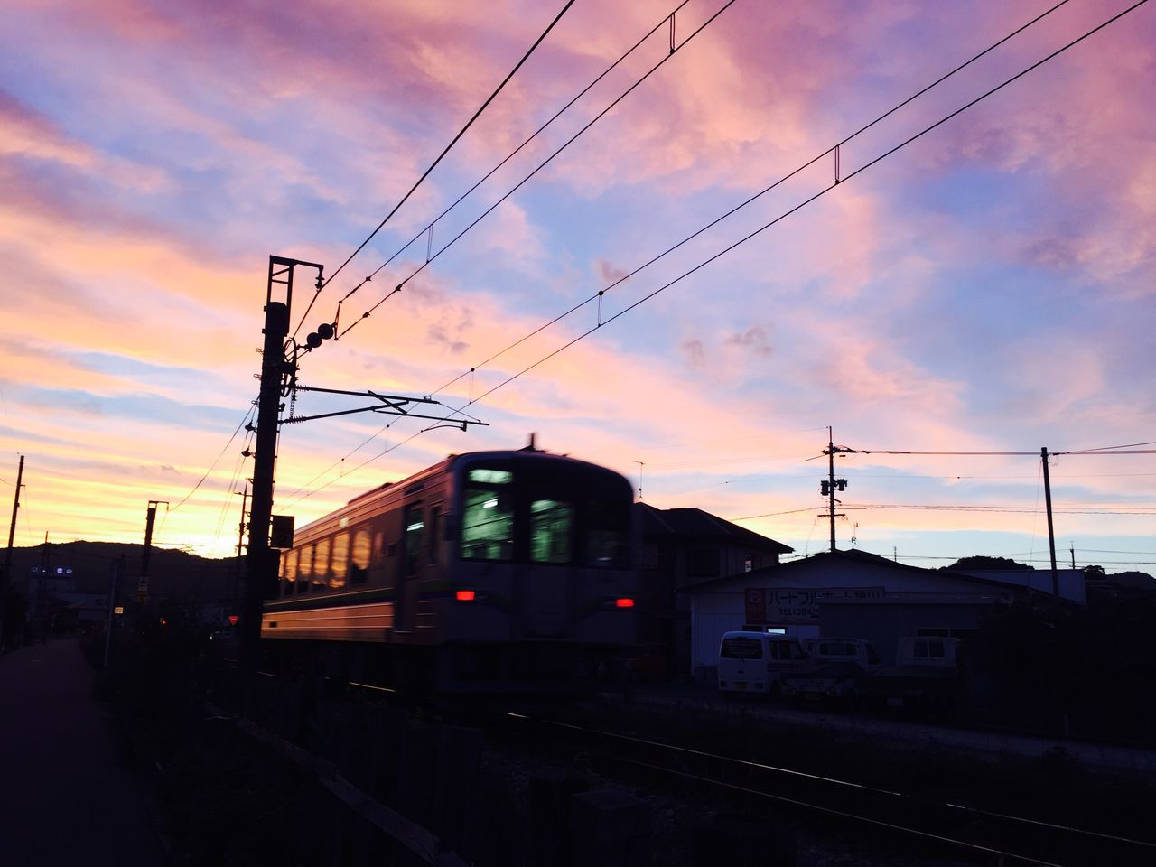 railroad track, transportation, rail transportation, sunset, public transportation, sky, power line, railroad station, cloud - sky, built structure, architecture, train - vehicle, railroad station platform, electricity pylon, mode of transport, cable, building exterior, connection, silhouette, orange color