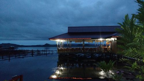 Illuminated building by sea against dramatic sky