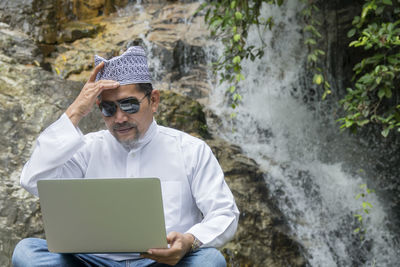 Mature man wearing sunglasses while using laptop against waterfall