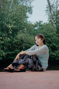 Young woman looking away while sitting on tree against plants