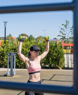 Happy woman in sportclothes working out with dumbbells on the sports ground in sunny summer day