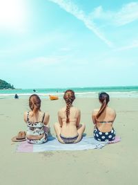 Rear view of female friends sitting on picnic blanket at beach against sky