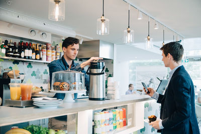 Businessman holding muffin and using phone while owner making coffee at office cafeteria