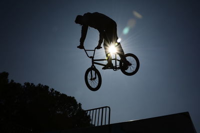 Low angle view of silhouette man doing stunt with bicycle against sky during sunny day