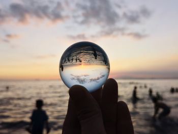 Close-up of hand holding crystal ball against sea and sky during sunset