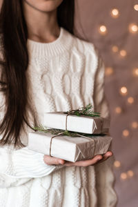 A girl in a white knitted sweater holds gifts in paper with a fir tree on a background of lights. 