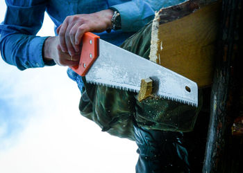 Low section of carpenter holding hand saw against sky