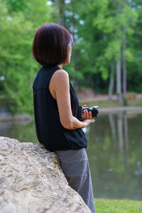 Side view of woman holding camera while leaning on rock