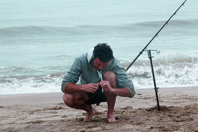 Full length of man crouching on beach with fishing rod