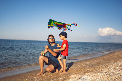 Father and son are standing on a sandy beach by the sea and launch a toy striped kite in the summer