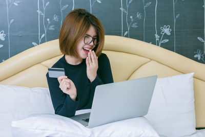 Smiling young woman doing online shopping over laptop while sitting on bed against wall at home