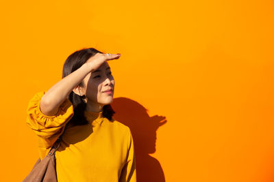 Woman shielding eyes while standing against yellow wall on sunny day