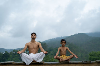 A young handsome boy doing yoga with a kid on the roof of a house situated in the middle of mountain