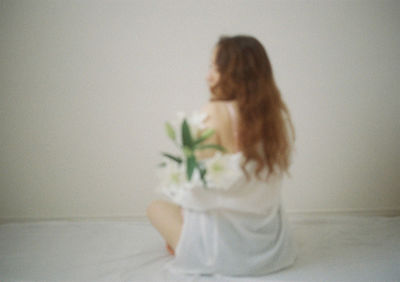 Rear view of woman sitting on bed against wall at home