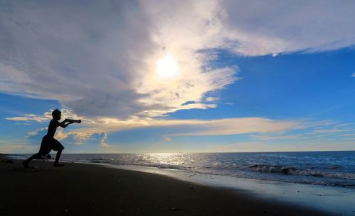 Full length of silhouette boy running at beach against cloudy sky