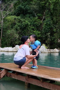 Woman kissing son on pier over lake against trees