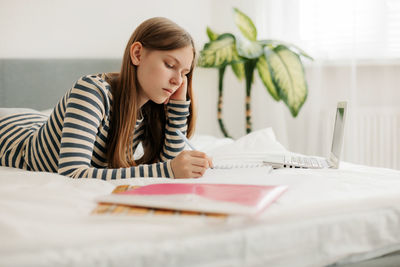 A charming teenage girl is lying on the bed, doing homework with a laptop, making notes 