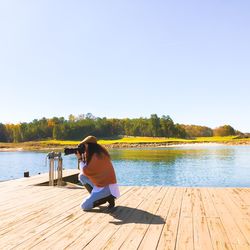 Woman photographing while kneeling on pier at lake against sky