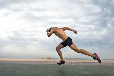 Side view of shirtless man taking running stance against sky