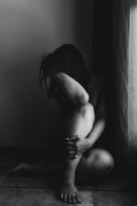 Side view of shirtless woman sitting against wall at home