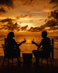Silhouette couple sitting on chairs by sea against sky during sunset