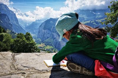Girl writing in book while sitting on rock against sky