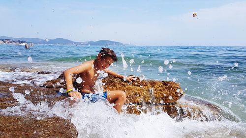 Happy shirtless boy sitting on rock with splashing wave in sea