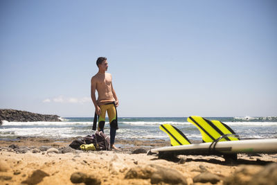 Full length of shirtless teenage boy standing at beach against clear sky