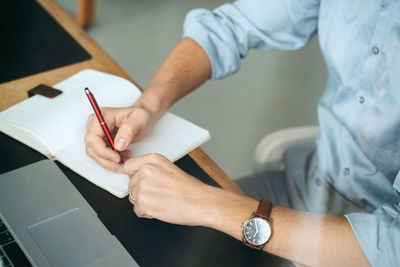 Unrecognizable crop male entrepreneur sitting at table and writing in notebook while planning business project