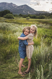 Portrait of happy sisters embracing while standing on grassy field in forest during sunset