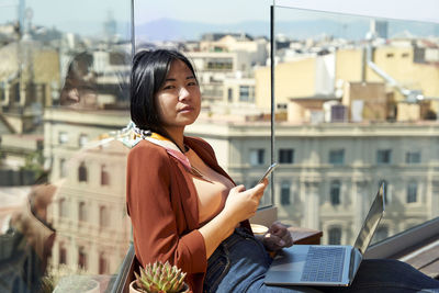 Beautiful woman with mobile phone and laptop leaning at terrace wall