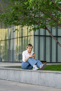Cute young woman sitting on bench in spring park under tree outdoors resting using smartphone