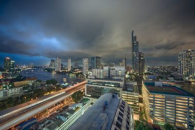Modern illuminated buildings and chao phraya river against cloudy sky at dusk