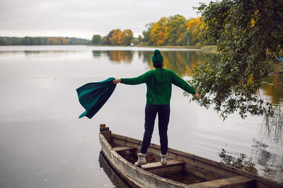 Woman in a green sweater stands with her back in an old wooden boat on a lake in autumn