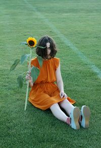 Full length of woman holding sunflower while sitting on grassy field