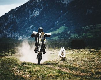 Dog chasing biker riding motorcycle on grassy landscape against mountain