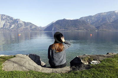 Rear view of woman wearing hat sitting at lakeshore against mountains