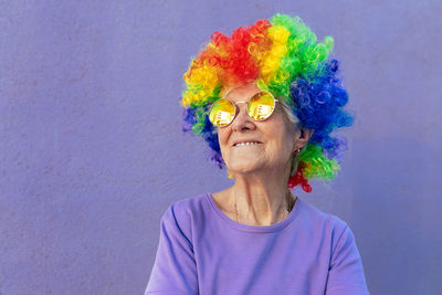 Content senior female athlete in modern sunglasses and bright wig standing on purple background